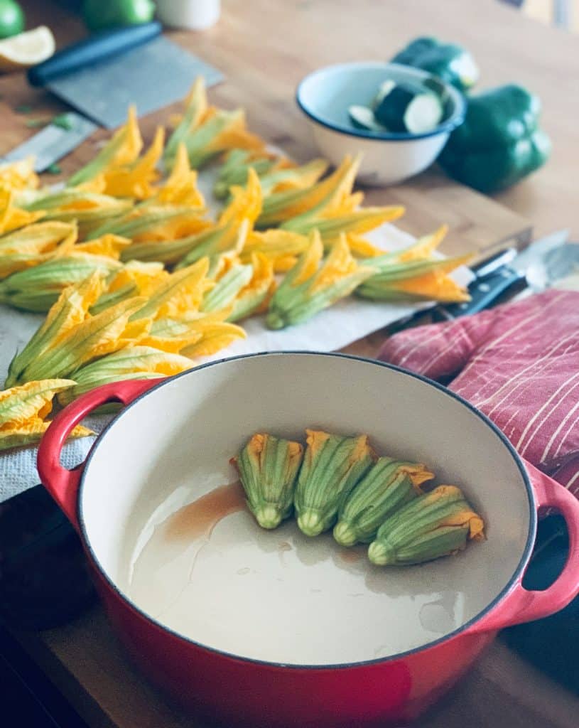 Stuffed squash blossoms in a red pot, in the background blossoms freshly washed on a paper towel and some cut up vegetables in a bowl.