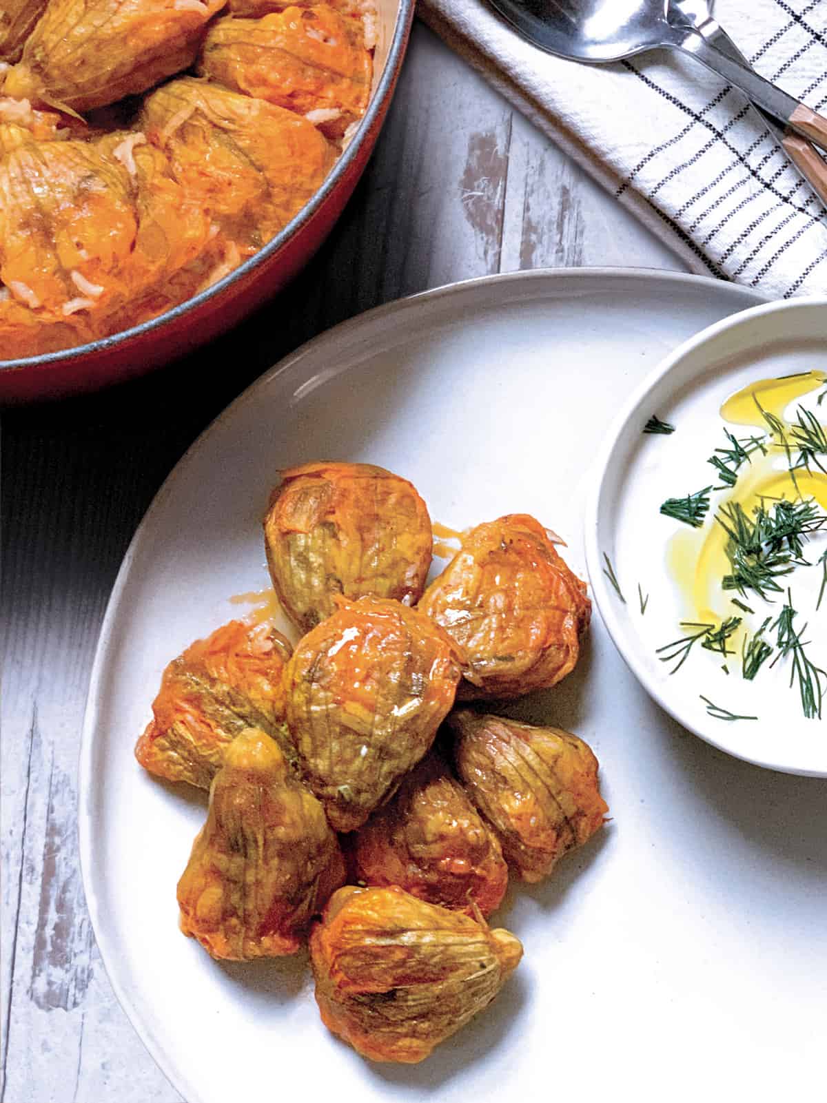A pot with zucchini flowers, a plate with stuffed flowers and a bowl with yogurt and herbs.