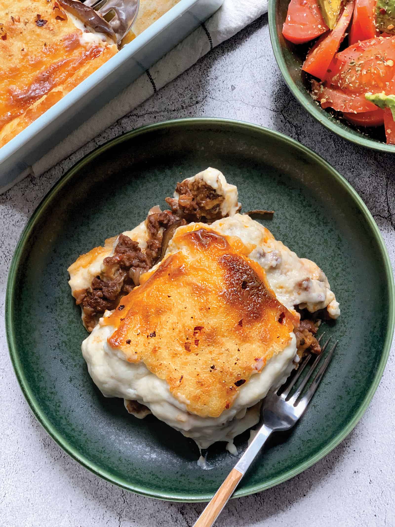 A green plate with a piece of vegetarian moussaka cooked with plant based meat Beyond beef, the baking pan with the moussaka and two serving spoons and a plate with a tomato/avocado salad.