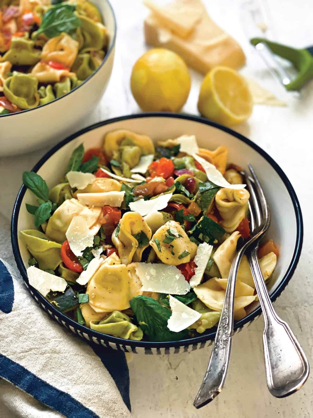 A bowl with pasta salad with zucchini, and tomatoes and a fork and spoon . At the back a bowl with salad, two lemons and a parmesan cheese block.