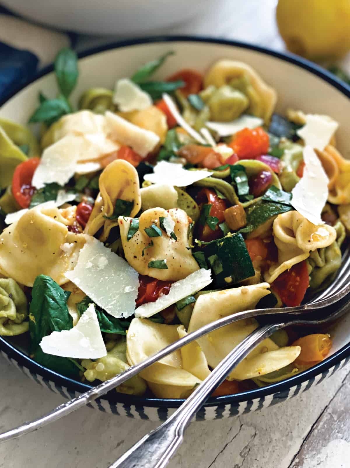  A plate with tortellini pasta salad with zucchini, and tomatoes and a fork and spoon inside.