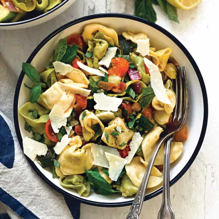 A white plate with colorful tortellini pasta with zucchini, tomatoes and shaved parmesan cheese.