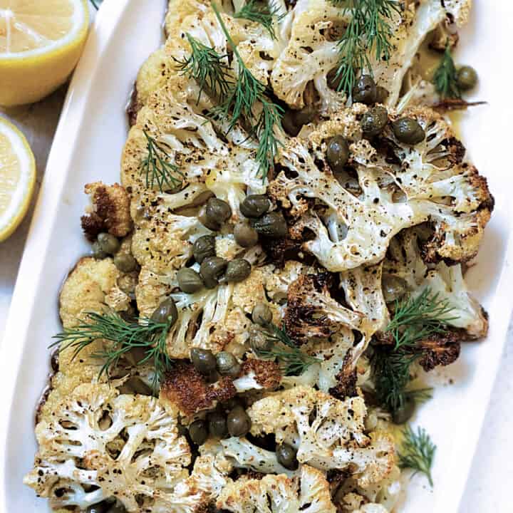 A serving platter with roasted cauliflower steaks in olive oil lemon and capers dressing, two lemon halves, a cloth napkin and some fresh dill leaves.