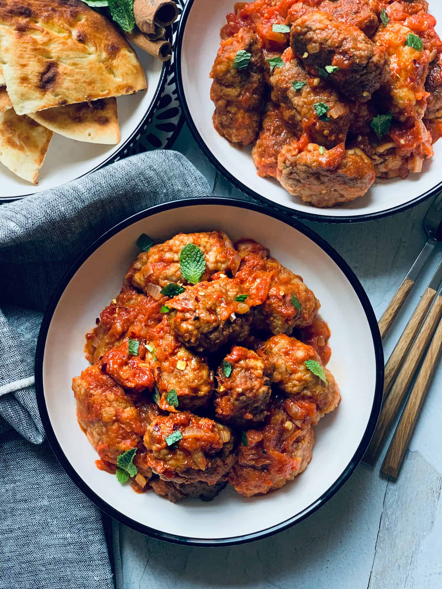 Two plates with cumin meatballs in red sauce-greek soutzoukakia and another plate in the back with cut up pita breads.