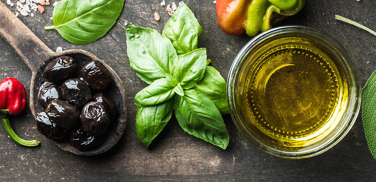 A wooden spoon filled with black olives, a small bunch of fresh basil, a glass jar with olive oil.