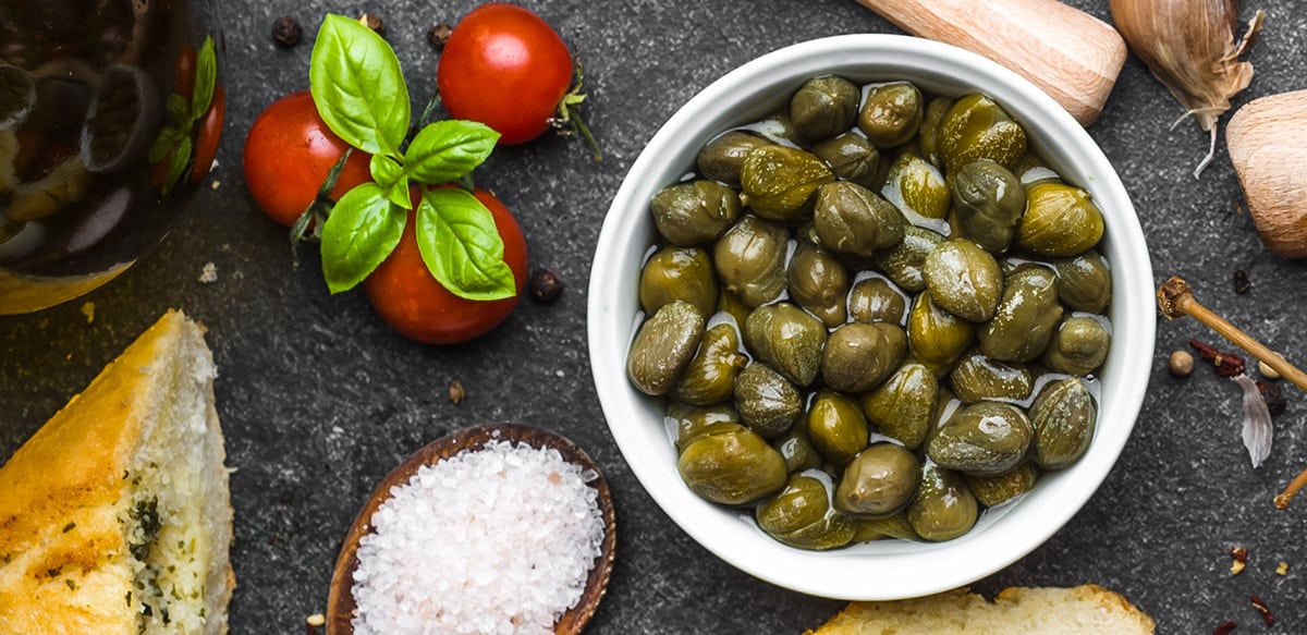 A bowl full of capers, 3-4 cherry tomatoes with fresh basil leaves on top slat and spices on a grey wood background.