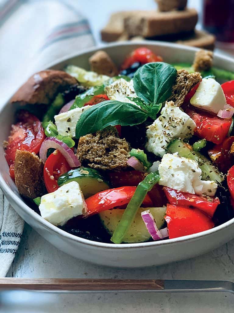 A Greek salad in a white plate with a serving spoon an olive oil bottle and fresh basil leave
