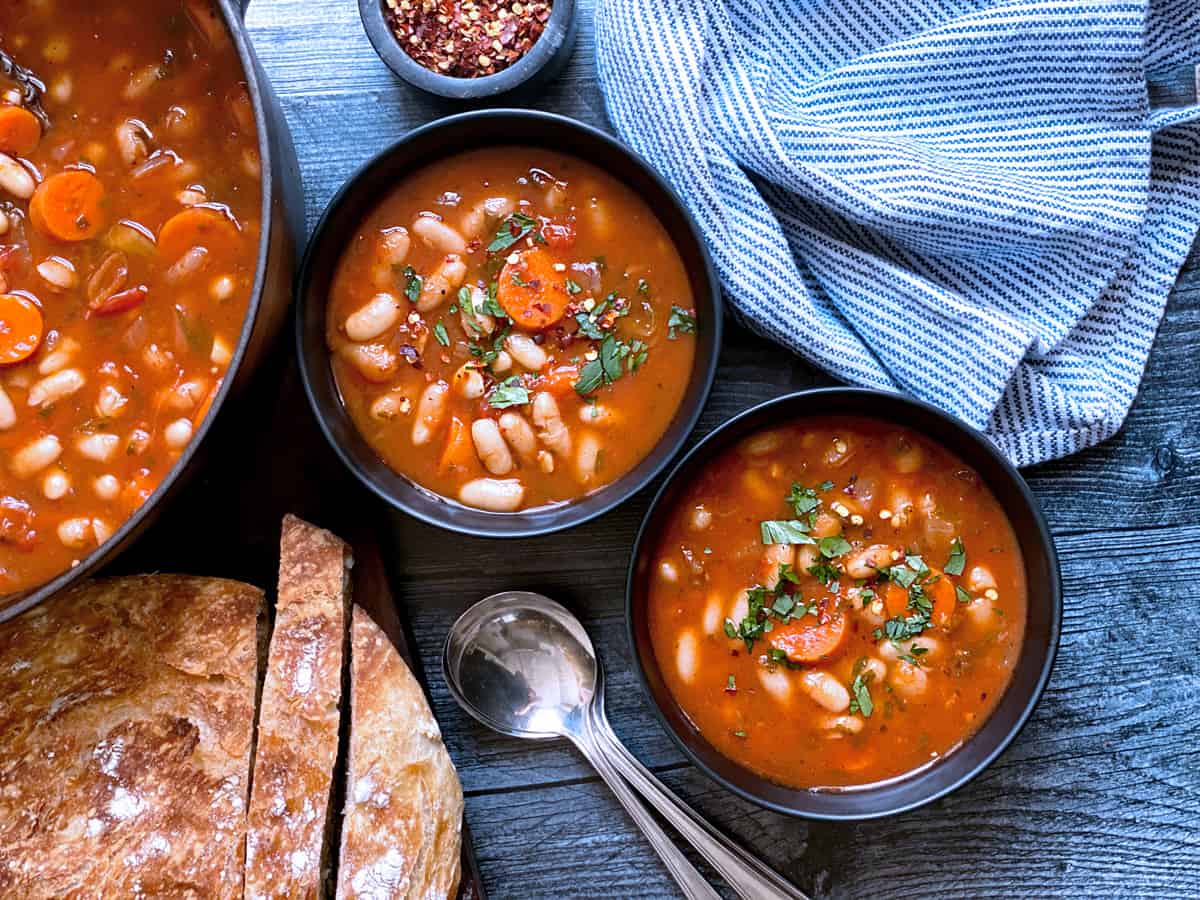 Two bowls with fasolada-greek white bean soup, bread and a pot filled with the soup.