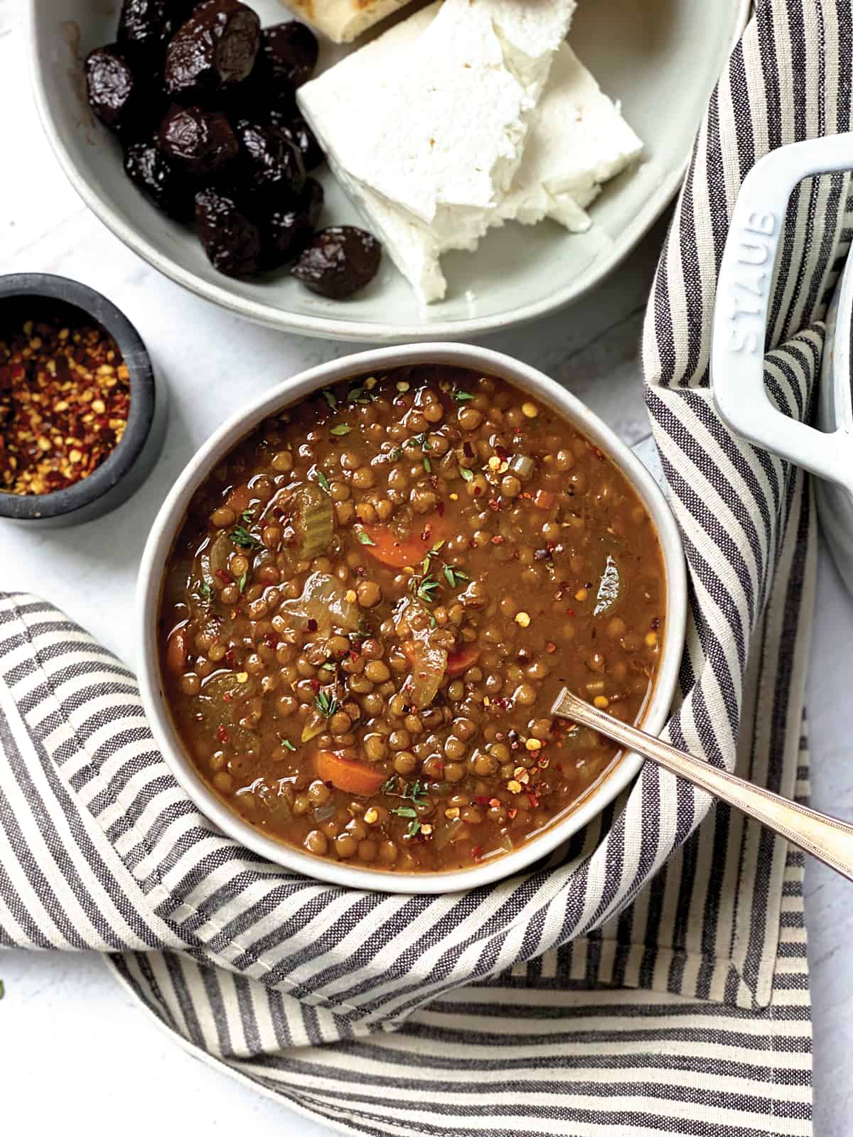 A bowl of greek lentil soup, a spoon,  a bowl with chili flakes and a plate with olives, feta and pitas and a napkin.