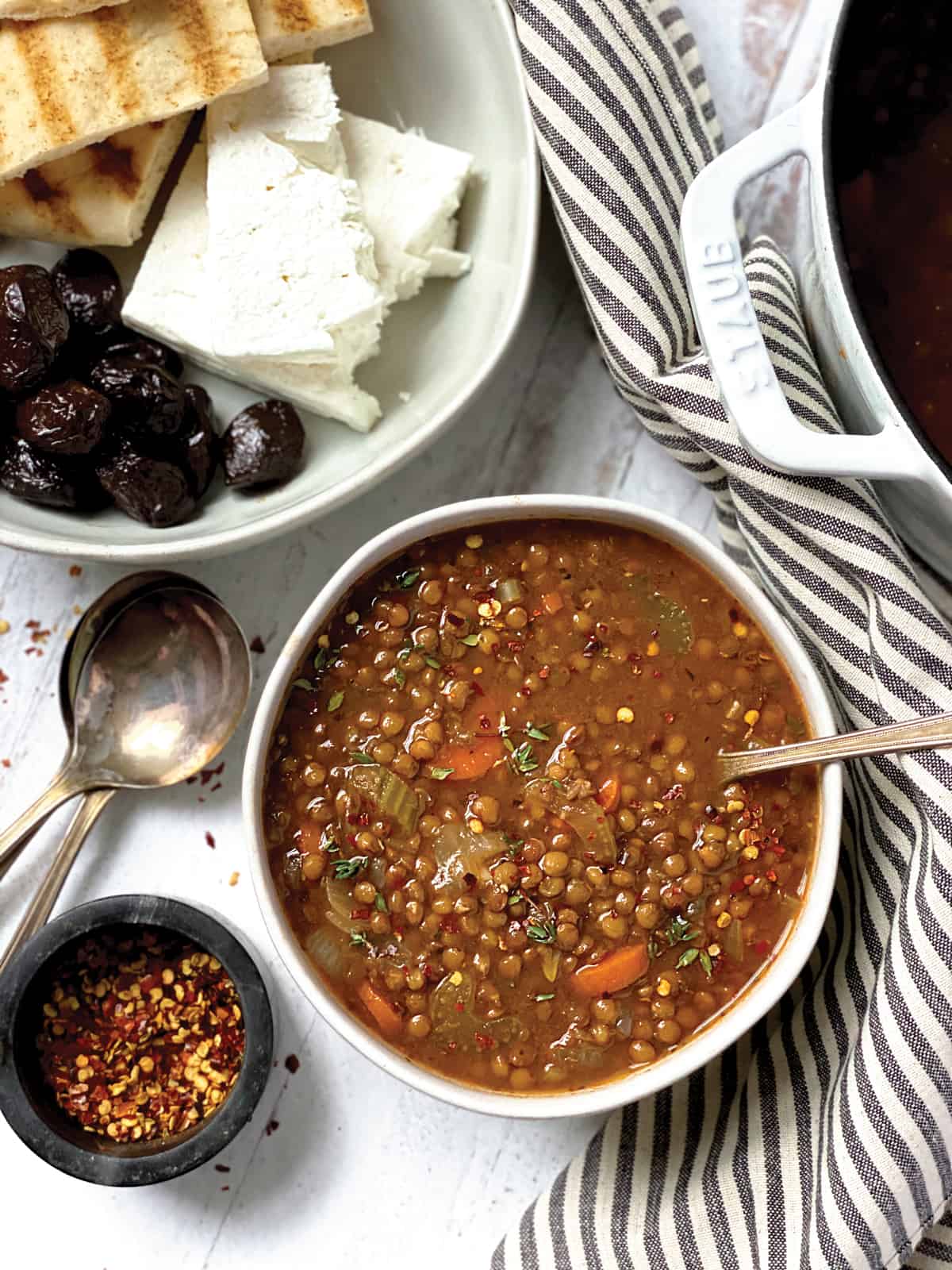 A bowl of greek lentil soup with a spoon, some spoons, a bowl of chili flakes and a plate with olives, feta and pitas at the back.