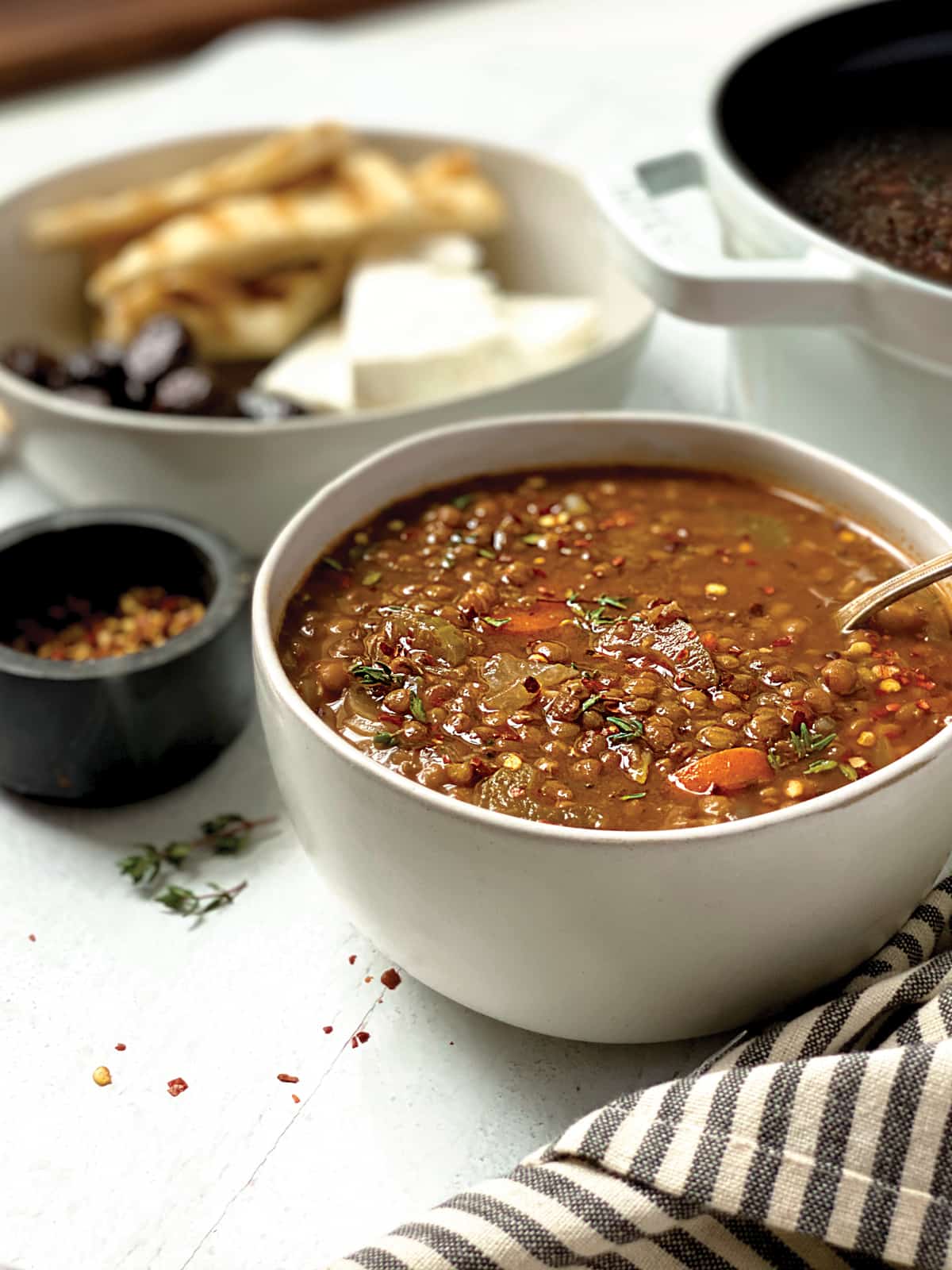 A bowl of greek lentil soup, a spoon, a plate with olives, feta and pitas at the back.