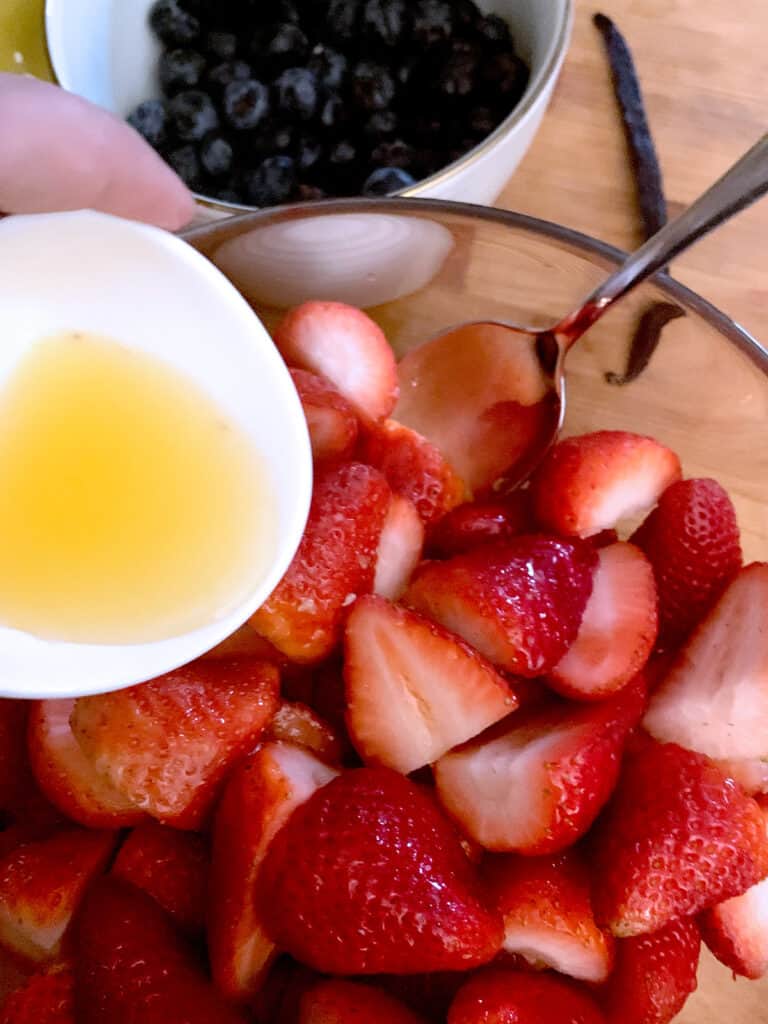 A bowl of strawberries, a hand pouring orange juice on them.