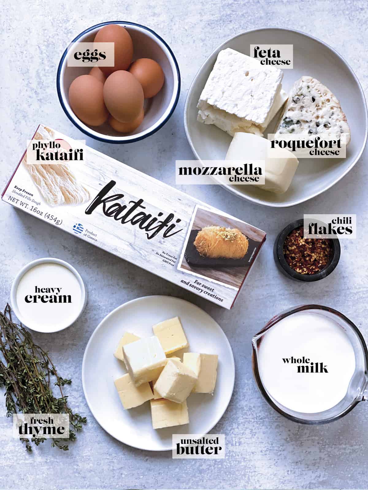 Ingredients for cheese pie. Eggs, a bowl with various cheese blocks, herbs, milk butter, phyllo pastry and cream.