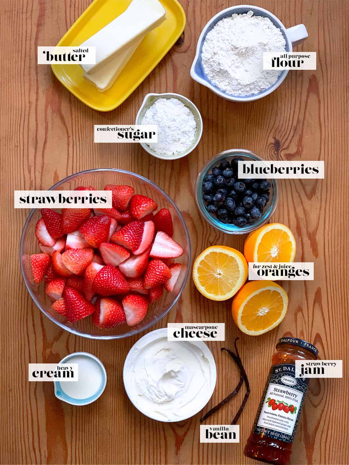 A plate with butter, oranges, a bowl with strawberries, a cup with sugar, a vanilla bean a cup of flour, and a jar of jam.