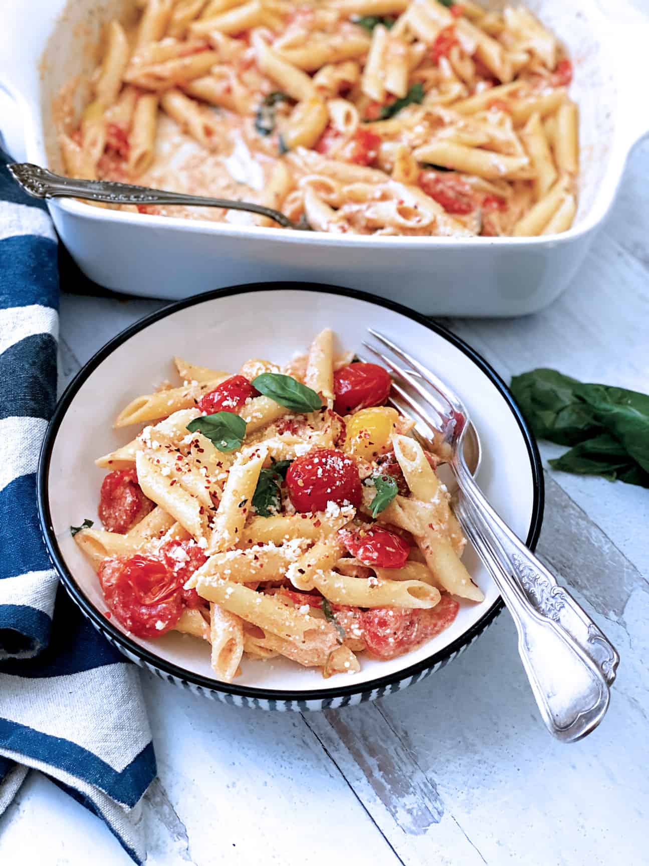 A plate with pasta with tomatoes and feta and fresh basil. A fork and spoon and a cloth napkin.