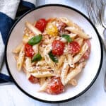 A plate with pasta with tomatoes and feta and fresh basil. A fork and spoon and a cloth napkin.