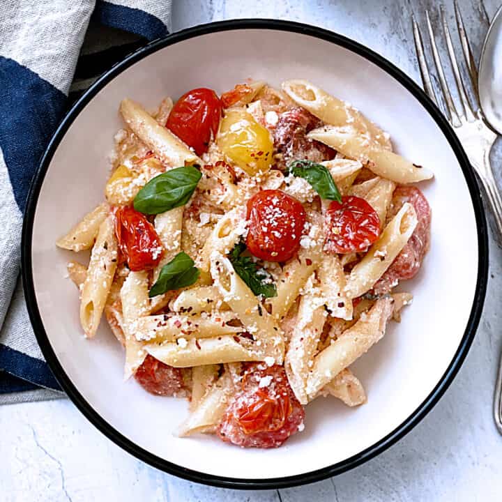A plate with pasta with tomatoes and feta and fresh basil. A fork and spoon and a cloth napkin.