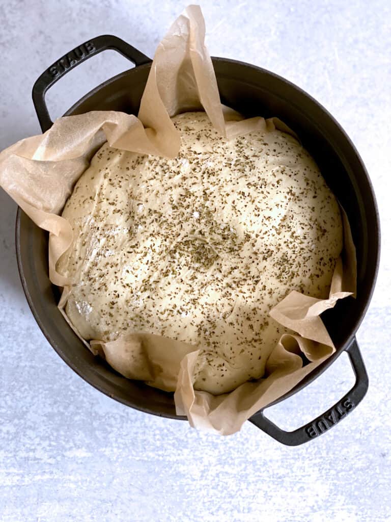 A black dutch oven with parchment paper and bread dough in it on a cement surface.