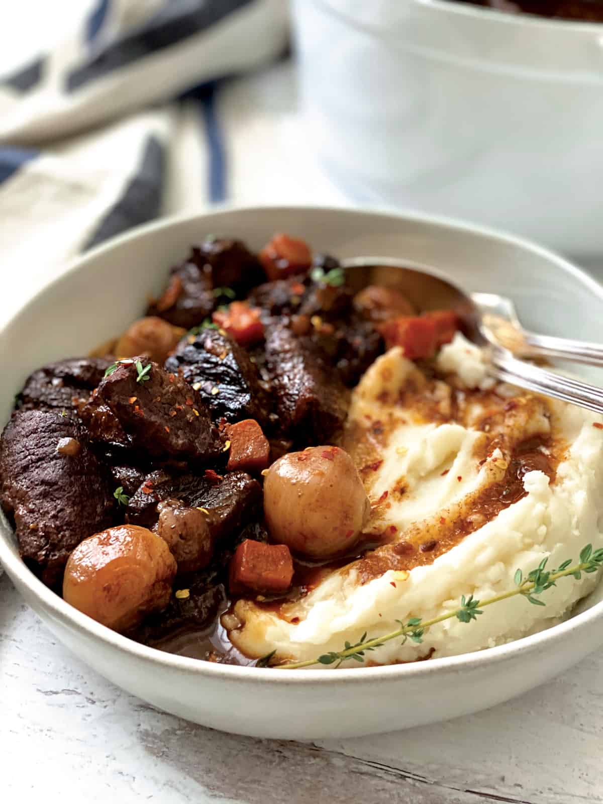 A plate with beef stew and mashed potatoes. At the back a white pot and a cloth napkin.