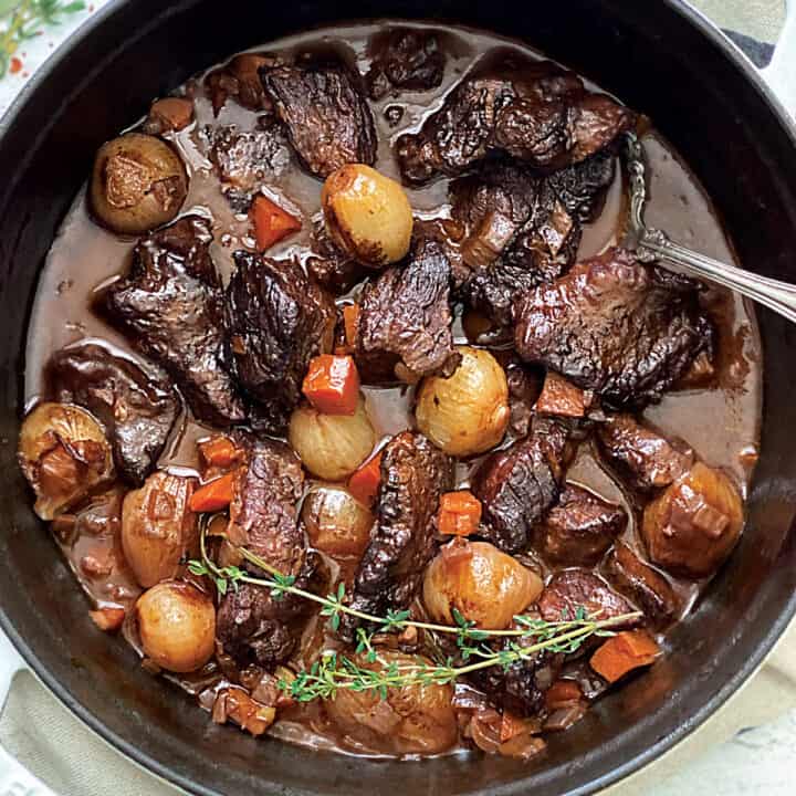 A white dutch oven with beef stew and a serving spoon on a cloth napkin.