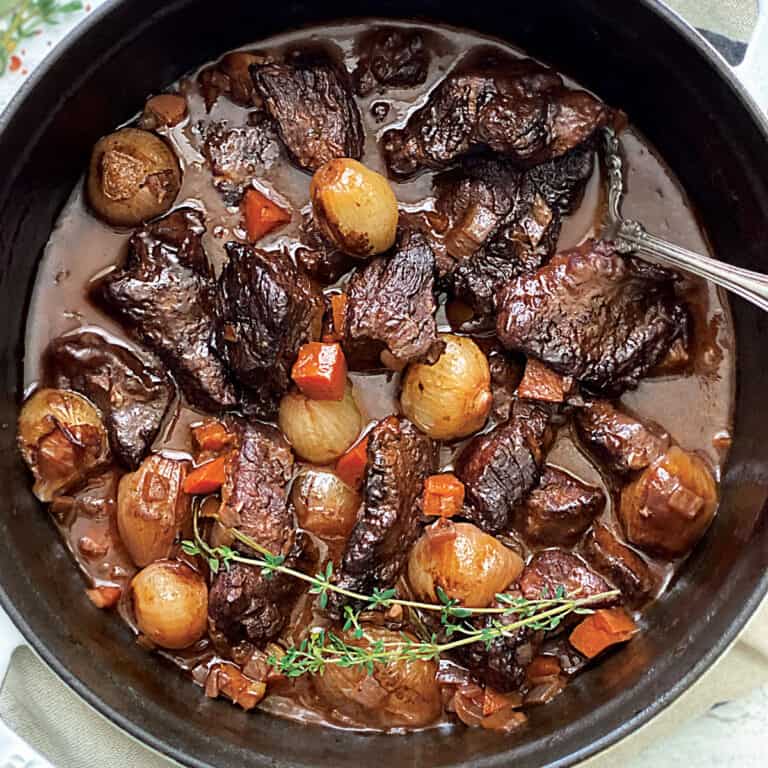 A white dutch oven with beef stew and a serving spoon on a cloth napkin.