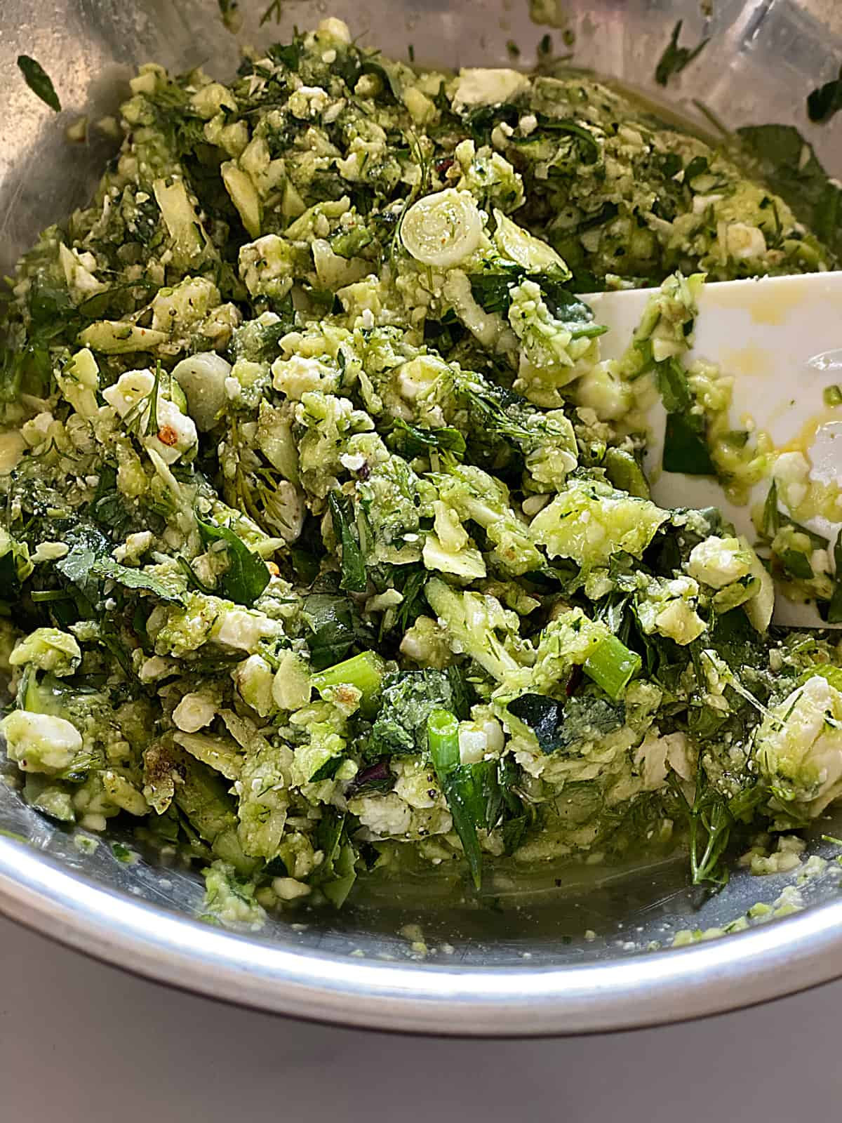 Grated zucchini with herbs and crumbled feta in a bowl with a white spoon.