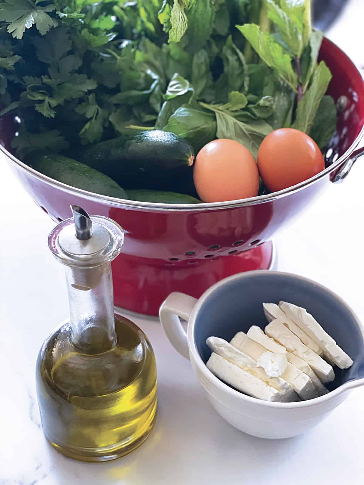 Fresh herbs, zucchini, eggs and scallions in a red colander. In front, a bottle with olive oil and a cup with feta cheese.