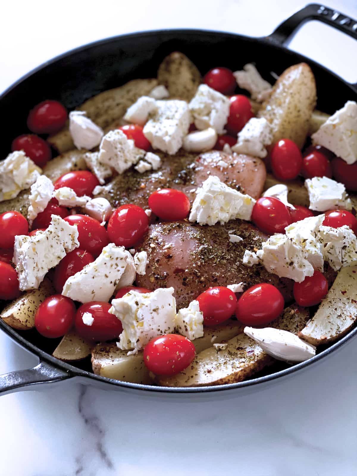 Chicken breast, cherry tomatoes, garlic cloves, potato wedges in a round black pan.