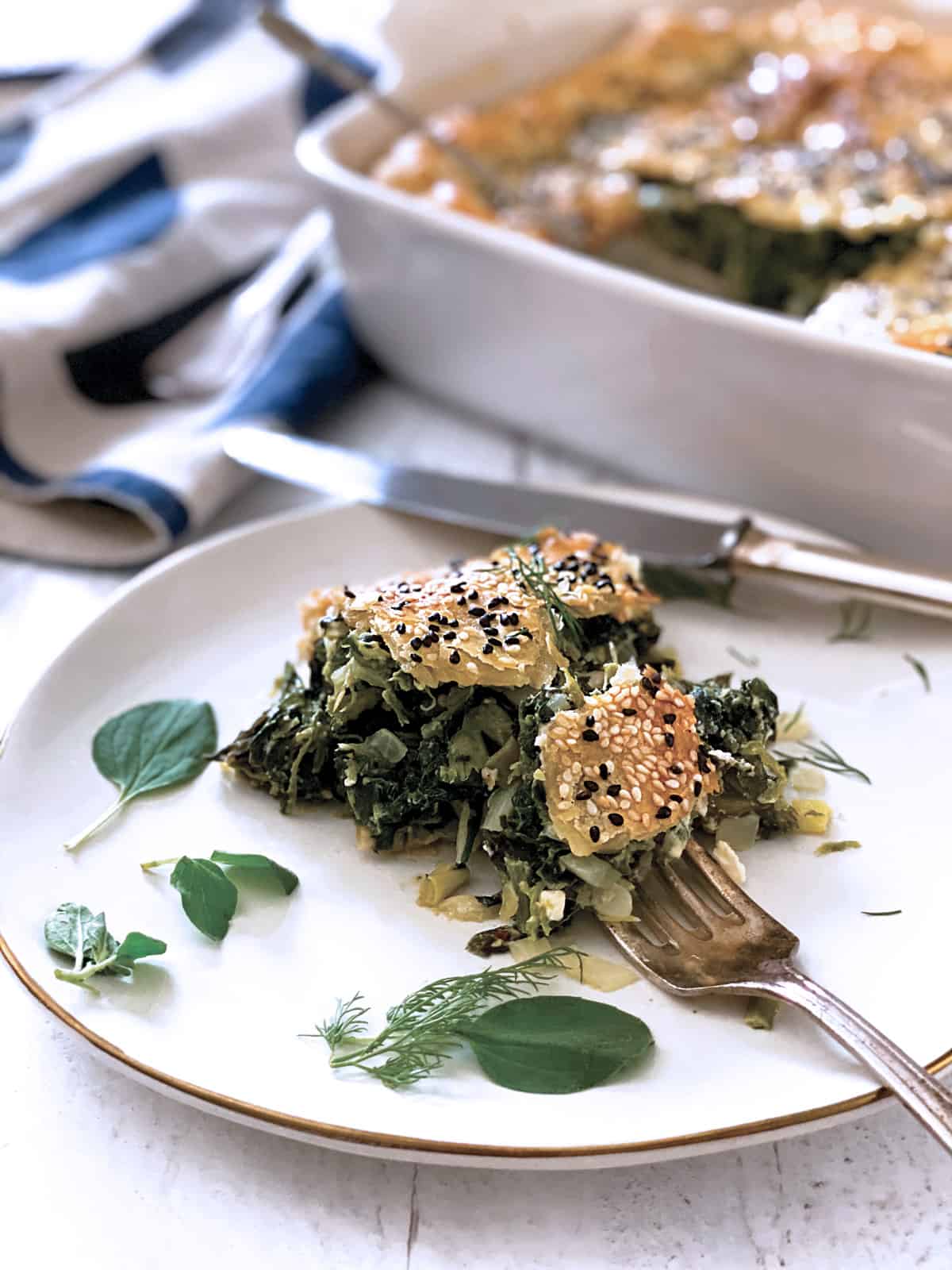 A piece of Greek Savory Pie in a white plate, a baking pan at the back, utencils and a cloth napkin.