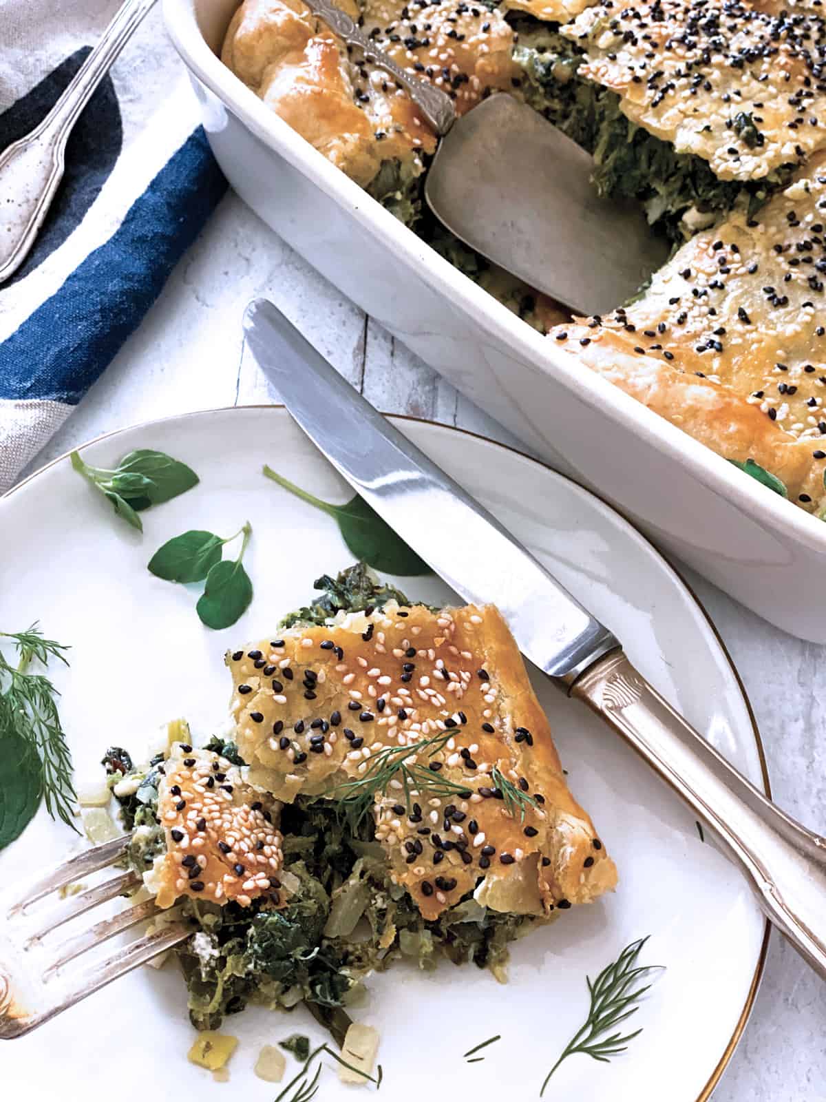 A piece of Greek Savory Pie in a white plate, a baking pan at the back, utencils and a cloth napkin.