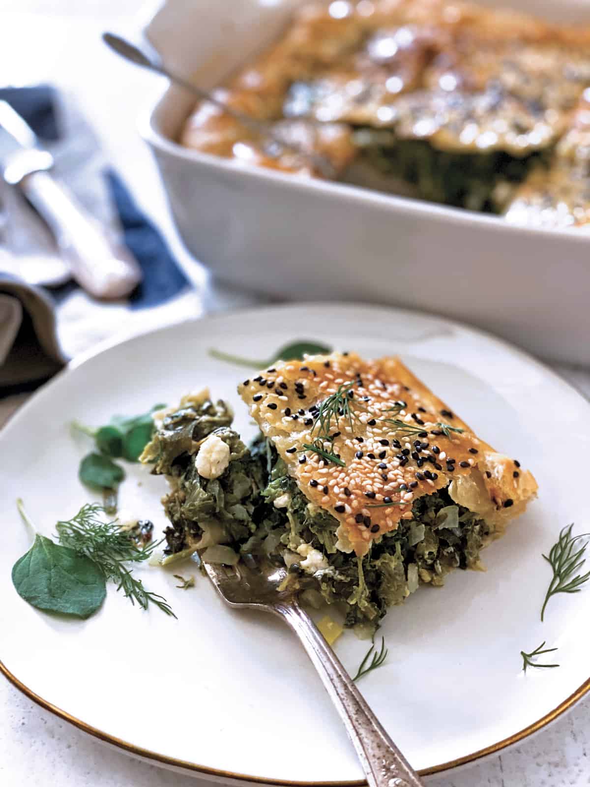 A piece of Greek Savory Pie in a white plate, a baking pan at the back, utencils and a cloth napkin.