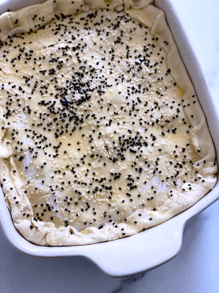 Uncooked pie with puff pastry and sesame seeds in a white baking pan.