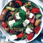 A Greek salad in a white plate with a serving spoon an olive oil bottle and fresh basil leave