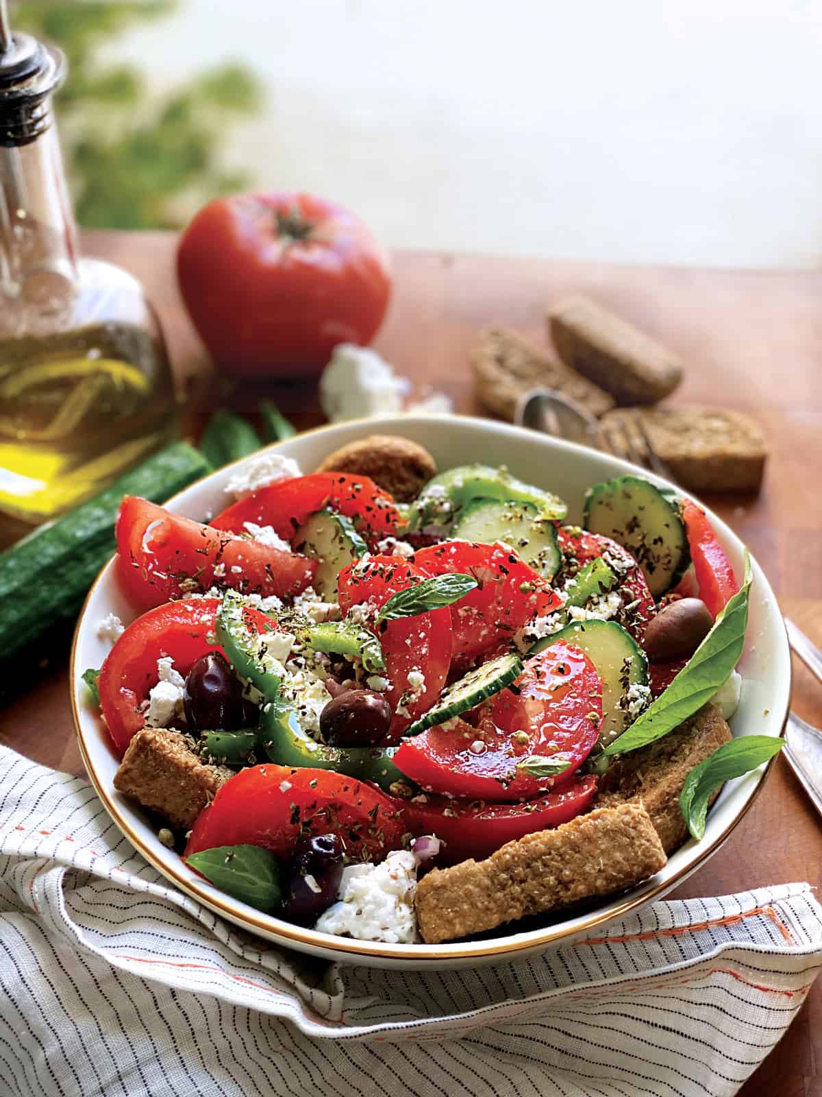 A plate with greek salad, a fork and a spoon an olive oil bottle, feta pieces and rusks, and fresh basil leaves.