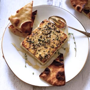 A block of roasted feta cheese with sesame seeds, honey and fresh thyme on a white plate with a spoon and cut up pita breads.