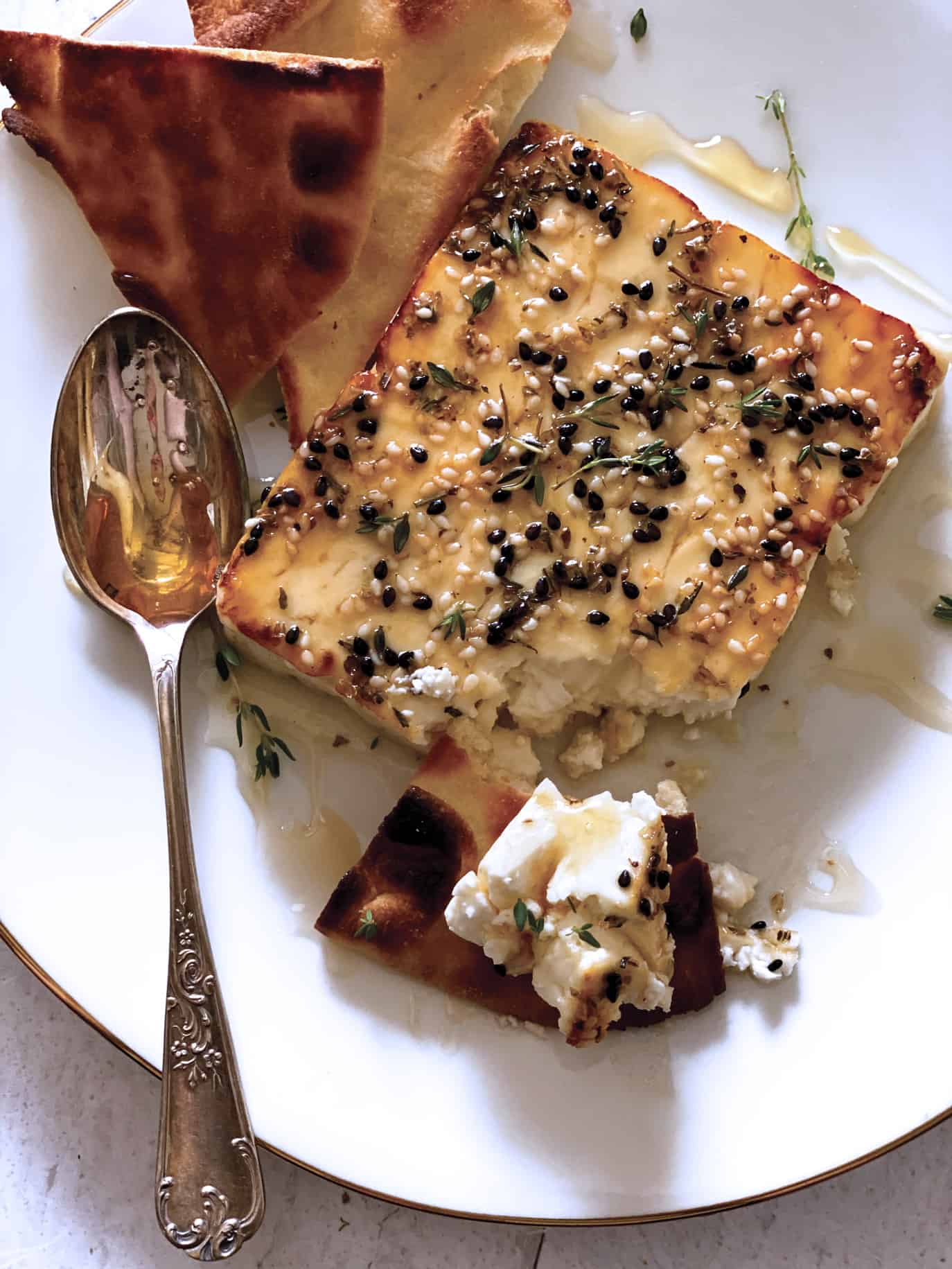 A block of roasted feta cheese with sesame seeds, honey and fresh thyme on a white plate with a spoon and cut up pita breads.