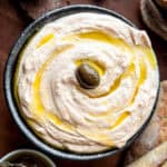 A bowl with greek whipped feta with an olive on top, a baguette and a bowl with olives in the front.
