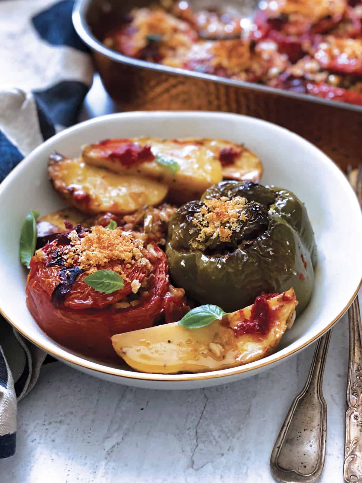 A plate with a stuffed tomato and a green bell pepper, potatoes and fresh herbs. A pan with food at the back and a cloth napkin.