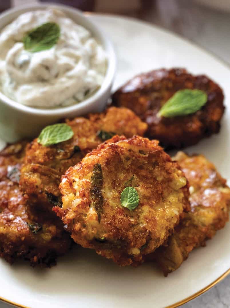 Four eggplant fritters on a plate with a container with yogurt dip, some fresh basil leaves and a cloth napkin at the back.