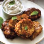 Four eggplant fritters on a plate with a container with yogurt dip, some fresh basil leaves and a cloth napkin at the back.