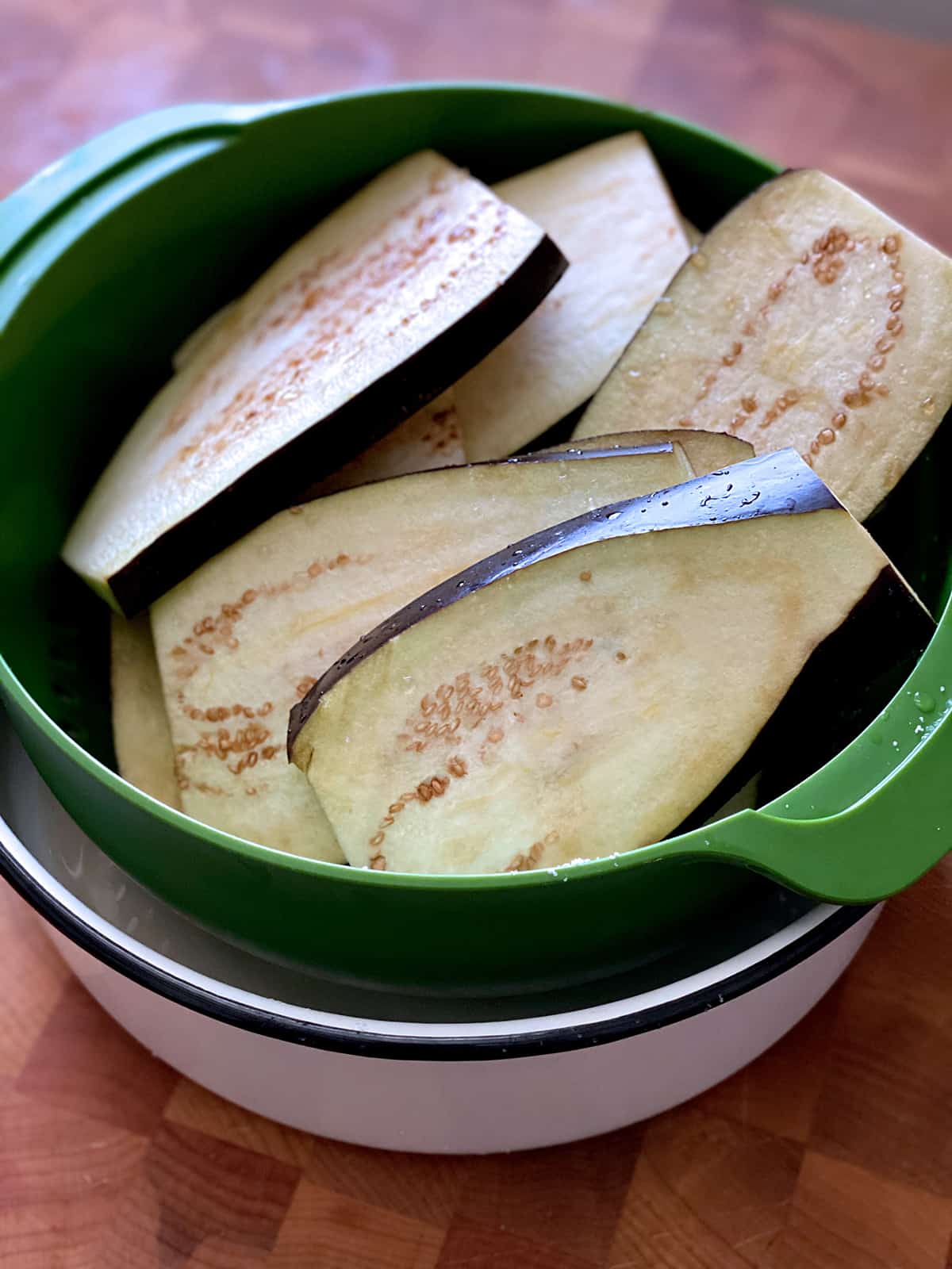 A green colander with eggplant slices in a white bowl on a butcher block.