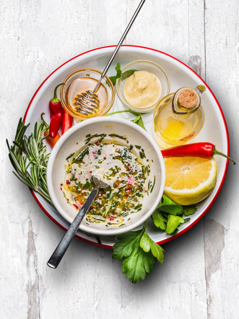 A plate with a bowl with herb dressing, a small ar with honey and a  wand, a glass bottle of olive oil, a small pot with mustard, half a lemon, red peppers, rosemary and parsley.