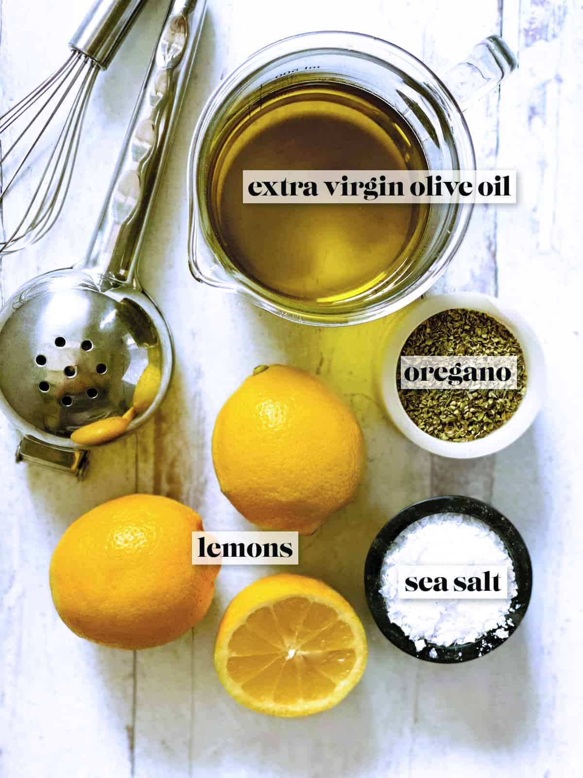 A glass jar with olive oil, a citrus juicer, a small whisk, 2 lemons and one half lemon, a pot with sea salt and another with dry oregano.