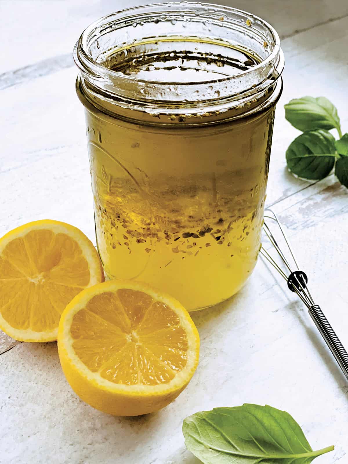 A glass jar with lemon and olive oil dressing-ladolemono, two lemon halves, a whisk and some fresh basil.