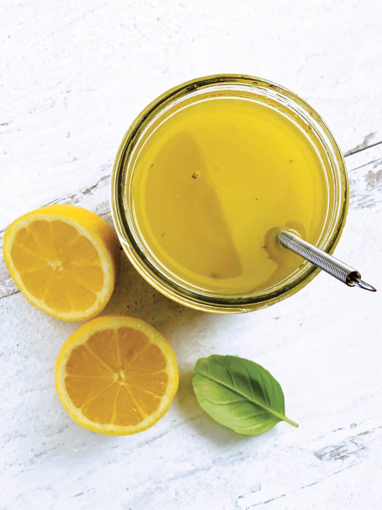 A glass jar with lemon and olive oil dressing-ladolemono, two lemon halves, a whisk and some fresh basil.