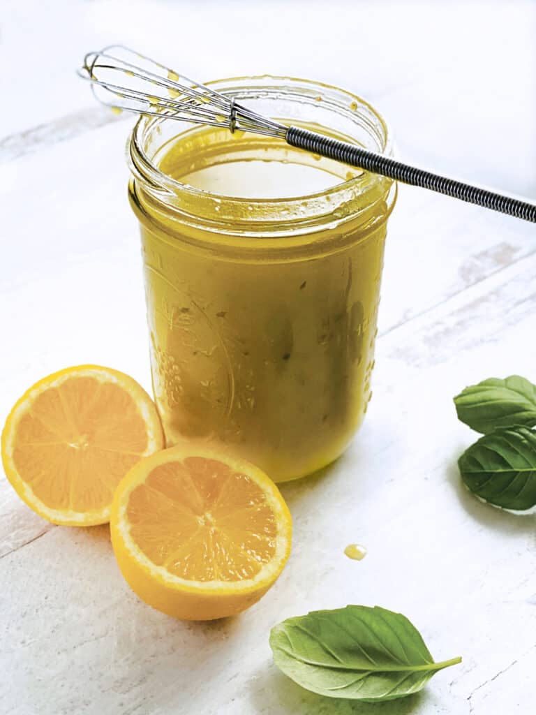A glass jar with lemon and olive oil dressing-ladolemono, two lemon halves, a whisk and some fresh basil.