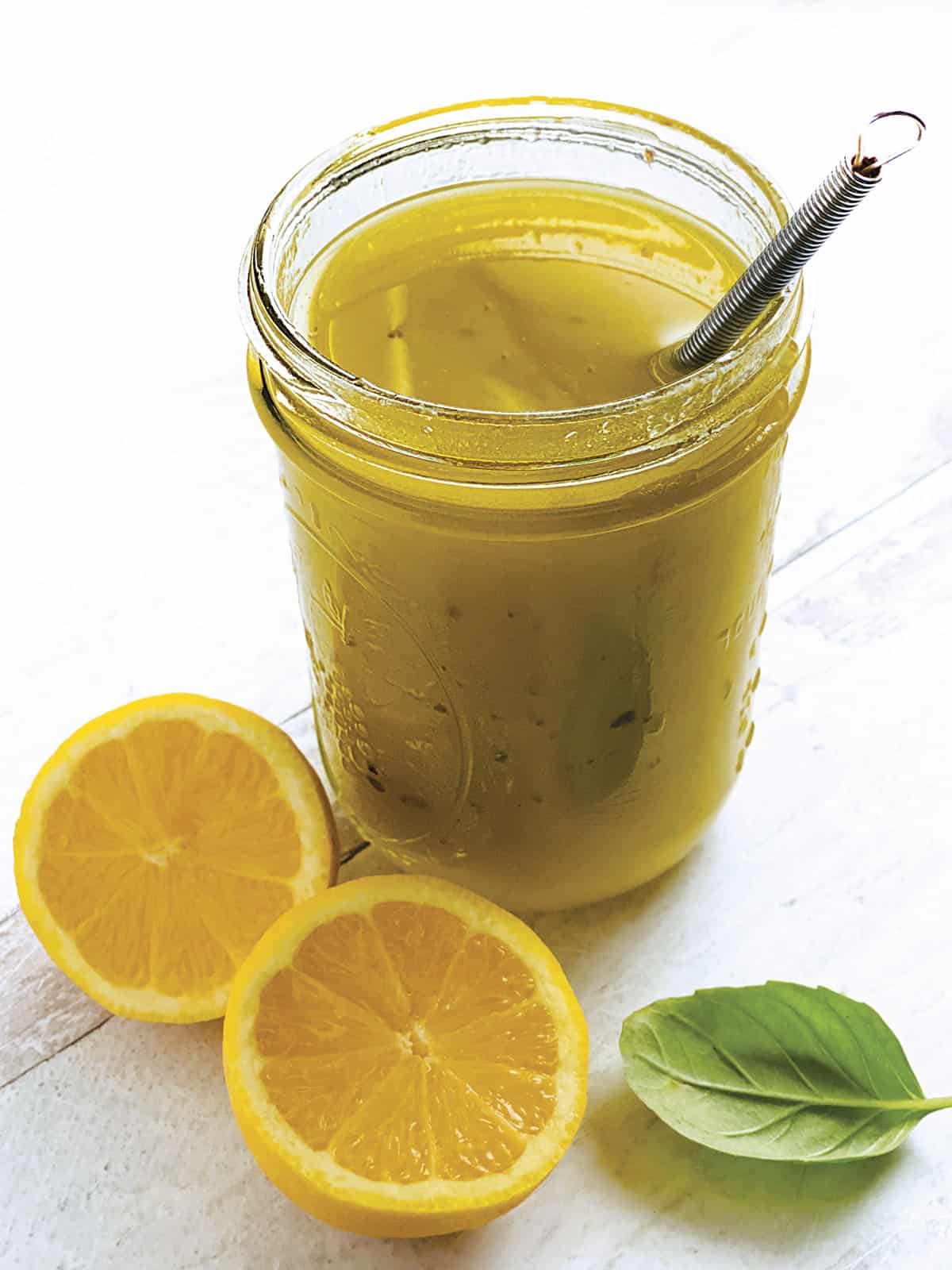 A glass jar with lemon and olive oil dressing-ladolemono, two lemon halves, a whisk and some fresh basil.