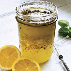 A glass jar with lemon and olive oil dressing-ladolemono, two lemon halves, a whisk and some fresh basil.