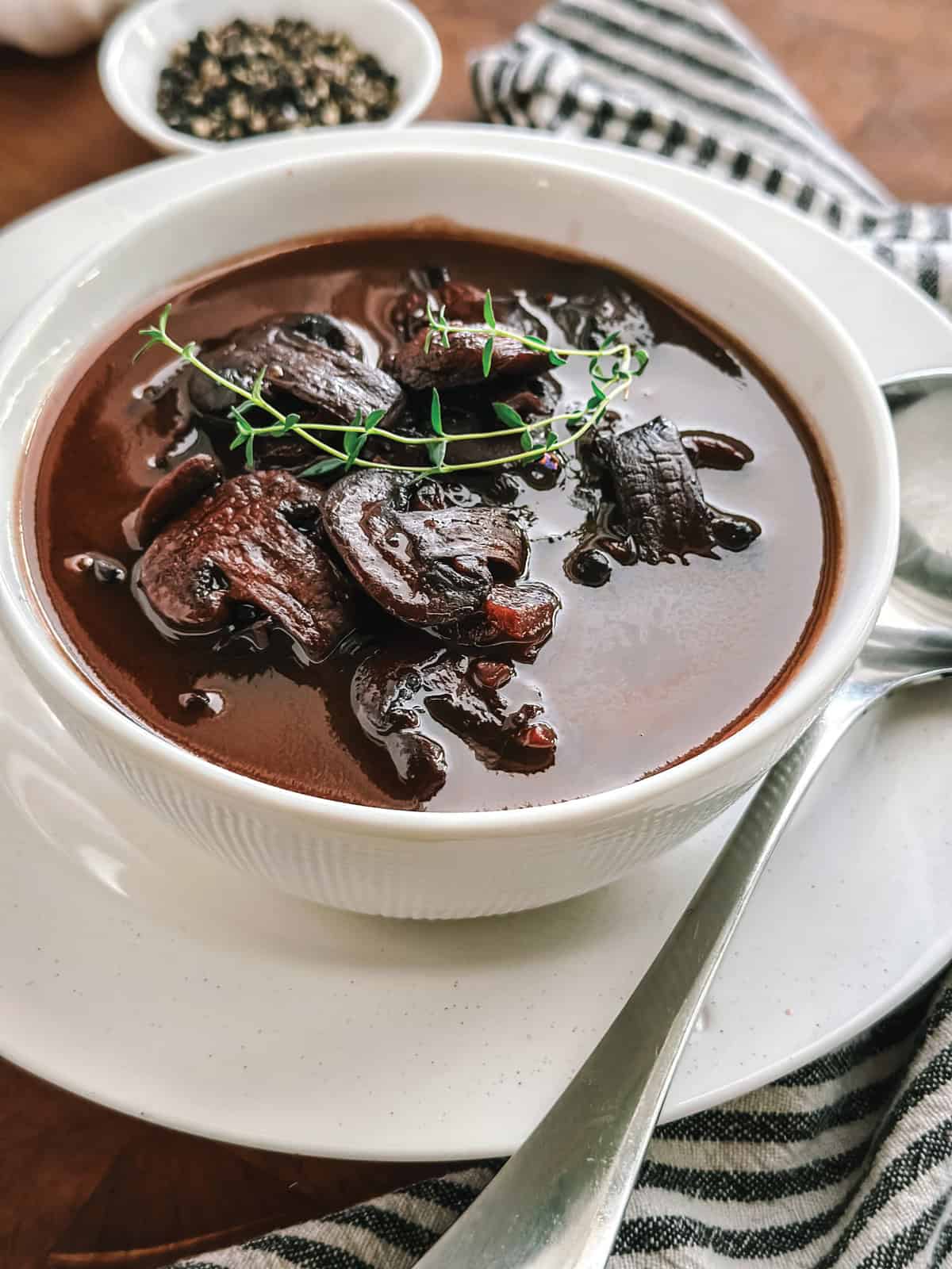 A bowl on a plate with marsalla sauce with mushrooms and twigs of thyme on top. A cloth napkin is under the plate, at the back, is a small container with pepper and sea salt.