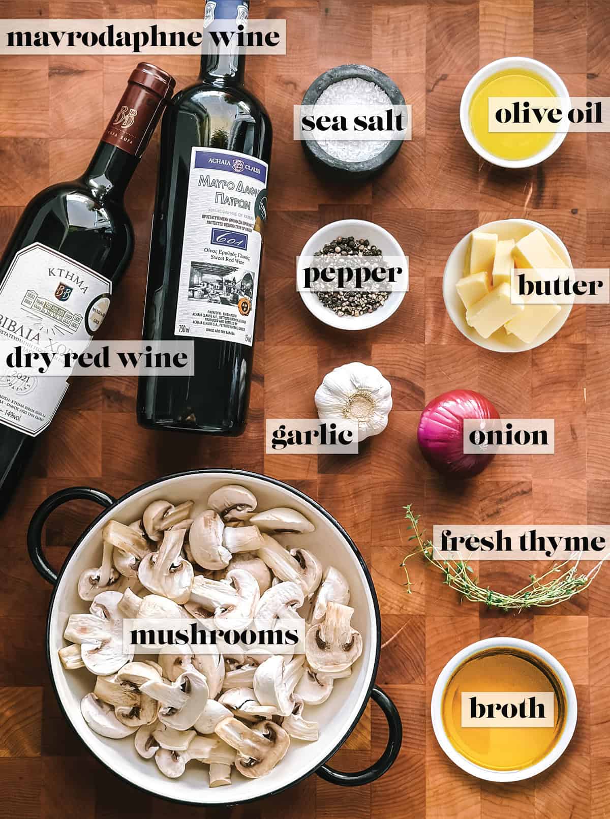 A bottle of dry red wine, a bottle of Mavrodaphne wine, a bowl with mushrooms, an onion, a garlic head, and small bowls with sea salt, pepper, butter, olive oil, broth and sprigs of fresh thyme on a wooden board.